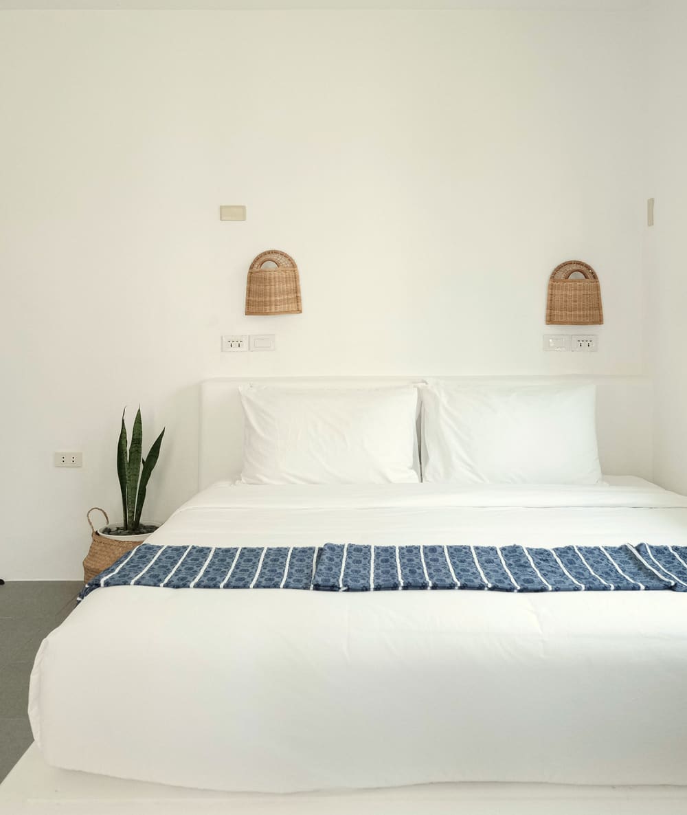 Double bed with white linen and a blue striped runner between two woven wall lamps, with a snake plant beside it.