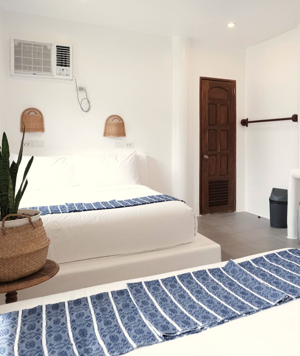 Room with two double beds with blue striped runners, a potted snake plant, woven wall lamps and a carved wooden door.