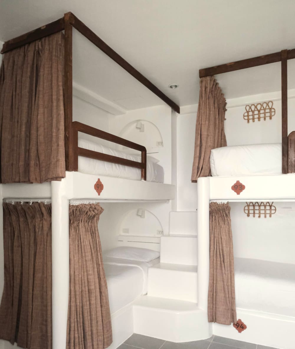 Dorm room with white built-in bunk beds, brown privacy curtains, wooden rails and a grey tiled floor.